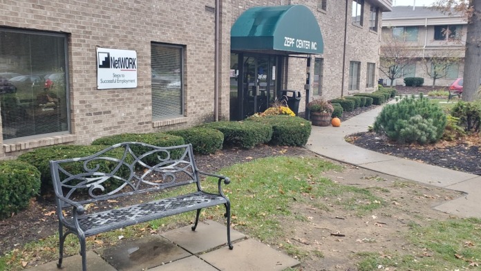 Exterior grounds showing metal bench seating, landscaped shrubs, walking path, flowering planters, and facility entrance with awning