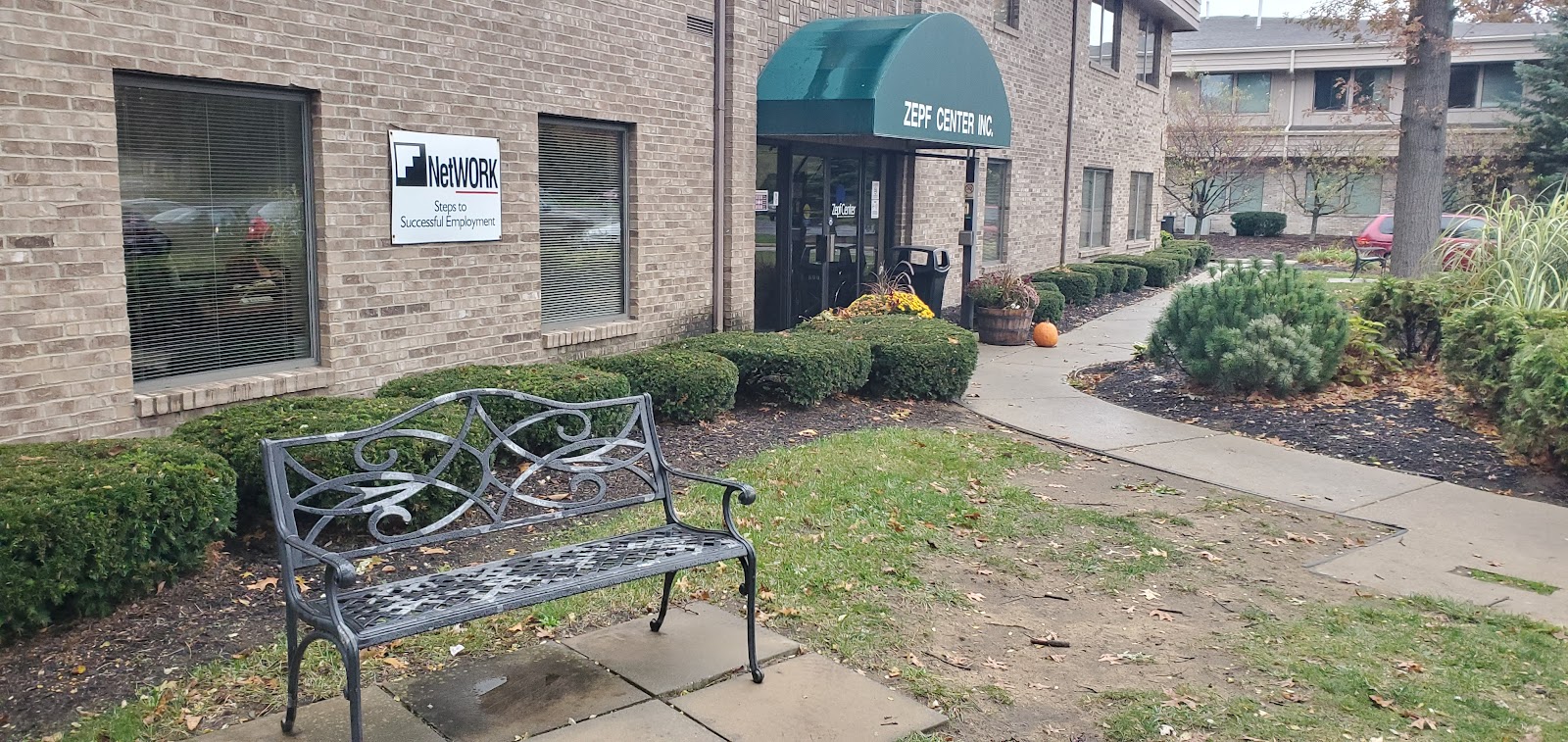 Exterior grounds showing metal bench seating, landscaped shrubs, walking path, flowering planters, and facility entrance with awning