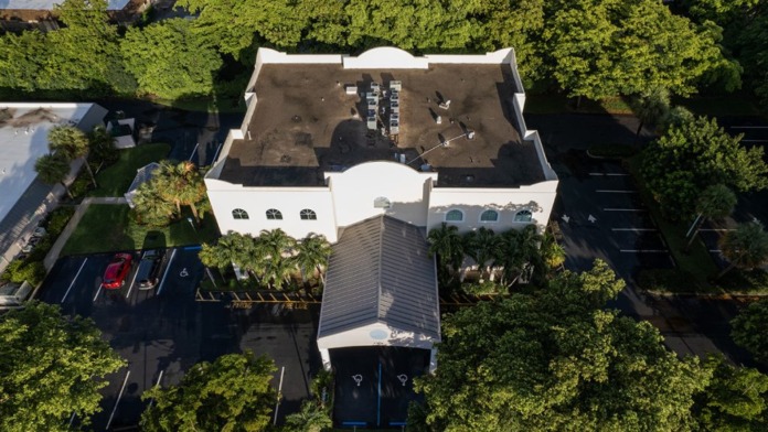 Aerial view of facility showing white building with courtyard, parking area, surrounding trees, and landscaping