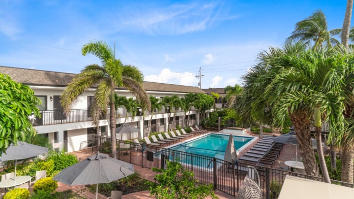Exterior courtyard featuring resort-style swimming pool, palm trees, multi-story white building with balconies, and lounge seating areas