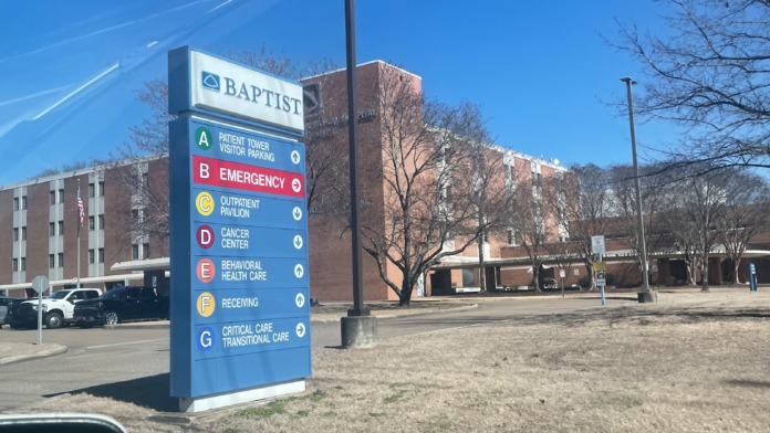 Exterior signage displaying Baptist hospital departments including emergency, outpatient, cancer center, and behavioral health care with parking area.