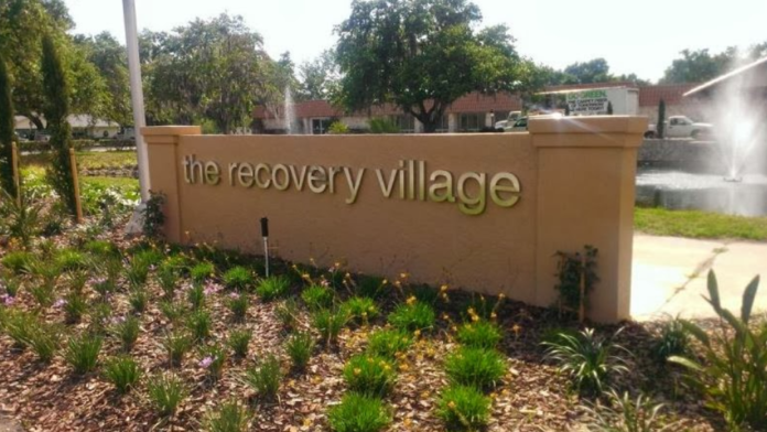 Exterior entrance sign reading 'the recovery village' with landscaped grounds and facility buildings in background