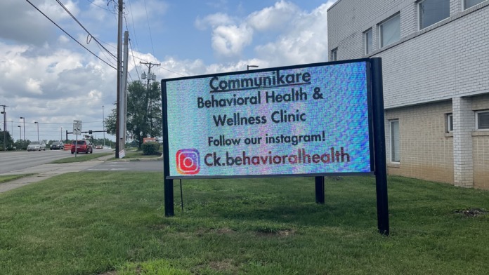 Outdoor signage for Communikare Behavioral Health & Wellness Clinic on grass with brick building and clear sky visible.
