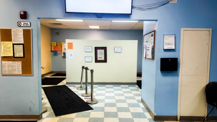 Interior entryway with checkered floor, blue walls, informational bulletin boards, rope barriers, mounted ceiling screen, and interior doors
