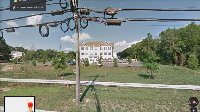 Exterior view of multi-story residential building with landscaped grounds, parking area, and surrounding vegetation