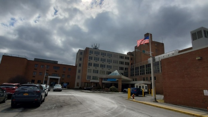 Exterior view of brick healthcare facility complex with multiple buildings, American flag, parking area, and cloudy sky