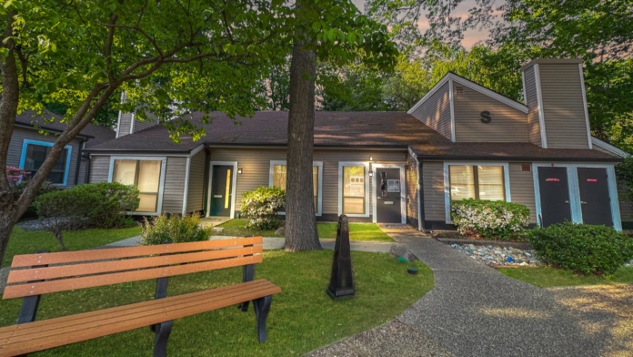 Exterior of treatment facility showing connected buildings with siding, mature trees, wooden seating areas, and manicured grounds