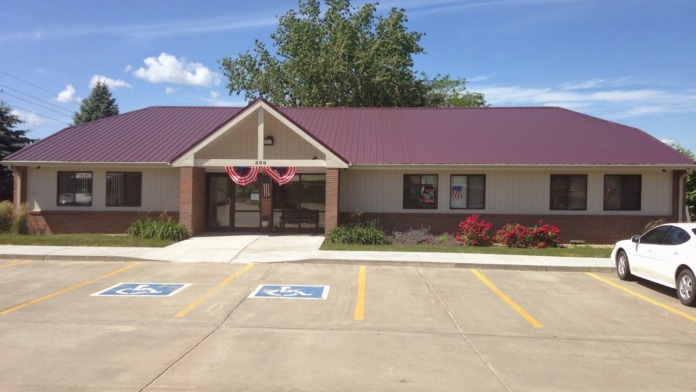 Exterior front of single-story facility building showing entrance, accessible parking spaces, landscaping, and patriotic bunting decorations.