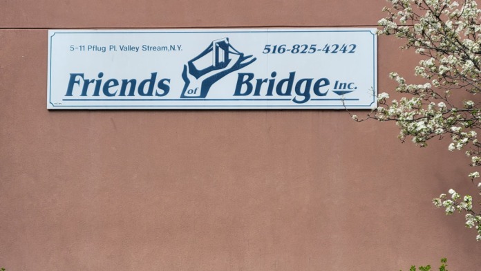 Exterior signage showing Friends of Bridge Inc. name, phone number, and address on brown wall with green landscaping