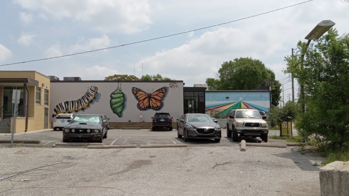 Exterior view of parking lot with multiple vehicles, street signage, mural artwork on building wall, and surrounding neighborhood