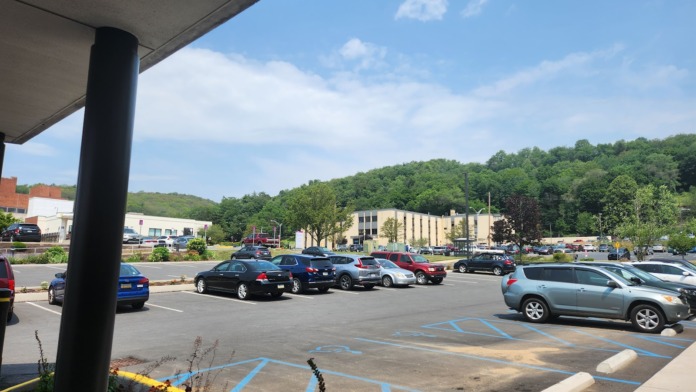 Exterior view of large parking lot with parked vehicles, landscaping, hillside background, and adjacent buildings