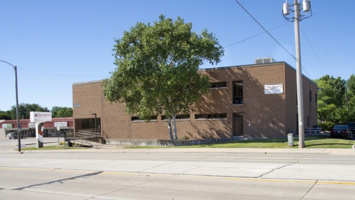 Exterior building view showing brick construction, large windows, entrance area, shade tree, and utility infrastructure.