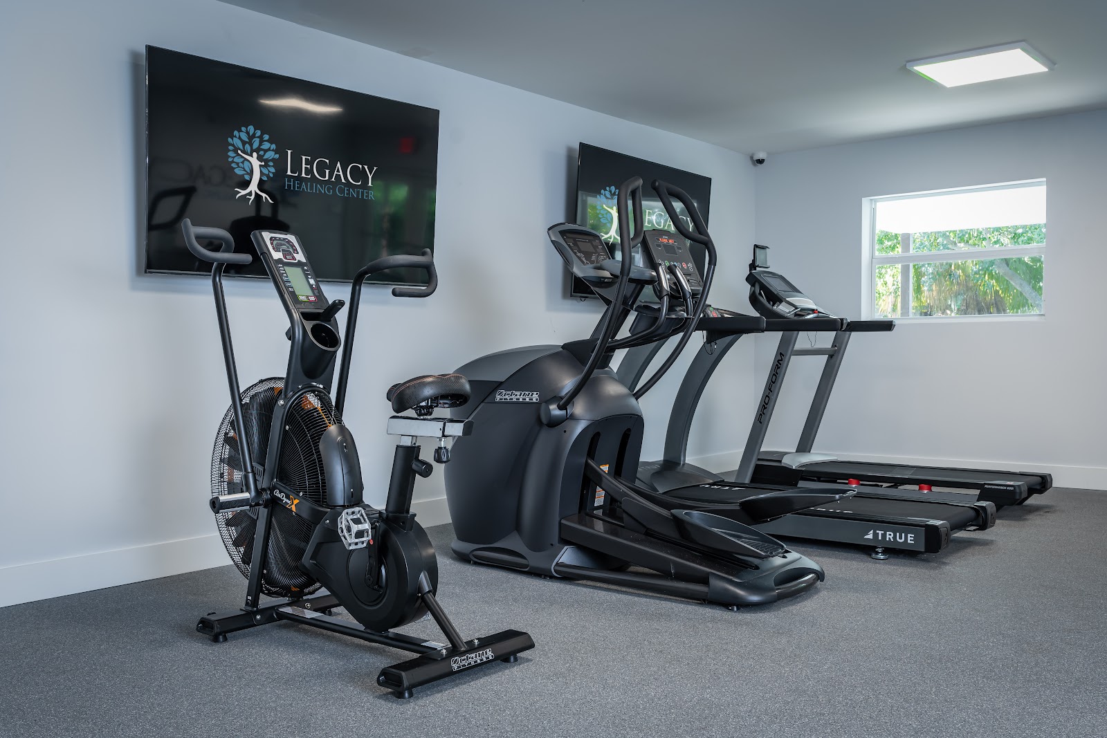 Interior fitness room with cardio equipment including stationary bike, elliptical machines, and treadmills with Legacy Healing Center branding on wall mounted monitor