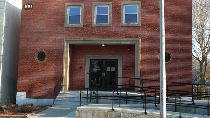 Front exterior of brick facility showing entrance portico, metal railings, and address number 100 on building face.