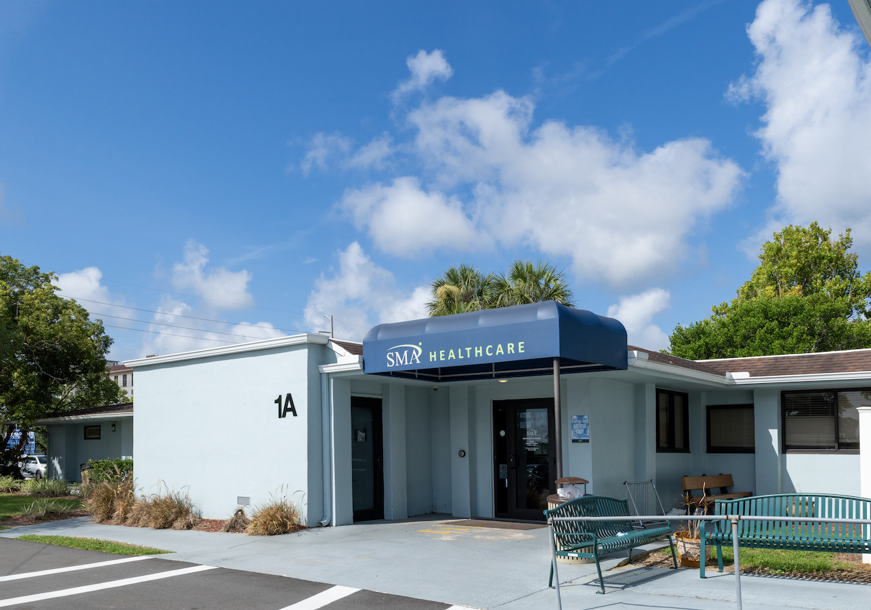 Exterior view of SMA Healthcare treatment center showing main entrance with blue awning, address 1A, landscaping, and outdoor seating area