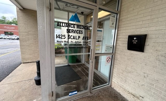 Exterior front entrance showing glass doors with Alliance Medical Services signage, brick building facade, and parking area.