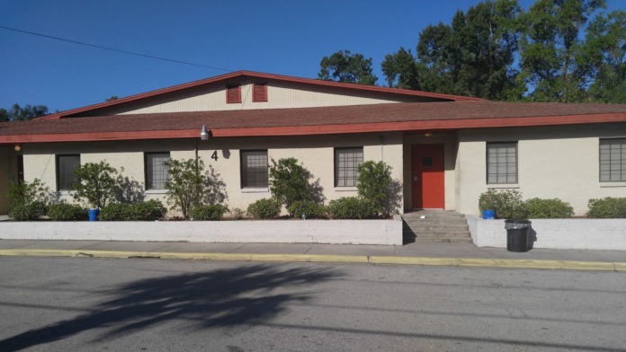 Exterior view of single-story treatment facility with white walls, red roof, red entrance door, and landscaping