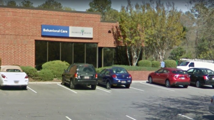 Exterior view of brick treatment facility with Behavioral Care signage, parking lot with vehicles, and landscaped grounds