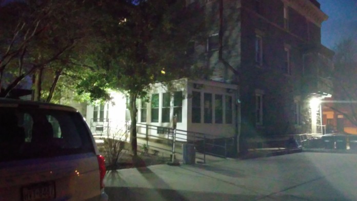 Exterior view of three-story brick recovery residence at dusk with illuminated entrance, mature landscaping, and street frontage.