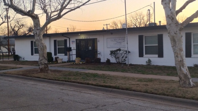 Exterior front view of white treatment facility with entrance, large trees, windows, and facility signage in evening light.