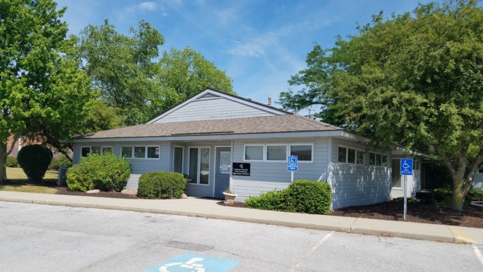 Exterior view of single-story treatment facility with gray siding, peaked roof, landscaping, and accessible parking spaces marked with blue wheelchair symbol.