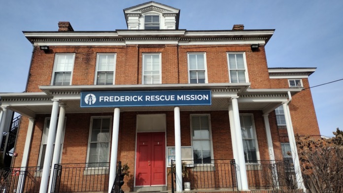 Exterior front view of historic brick facility with white columns, dormers, red doors, brick walkway, and mature landscaping