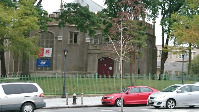 Exterior front facade showing stone building with banners and green fencing, visible from street with parked vehicles