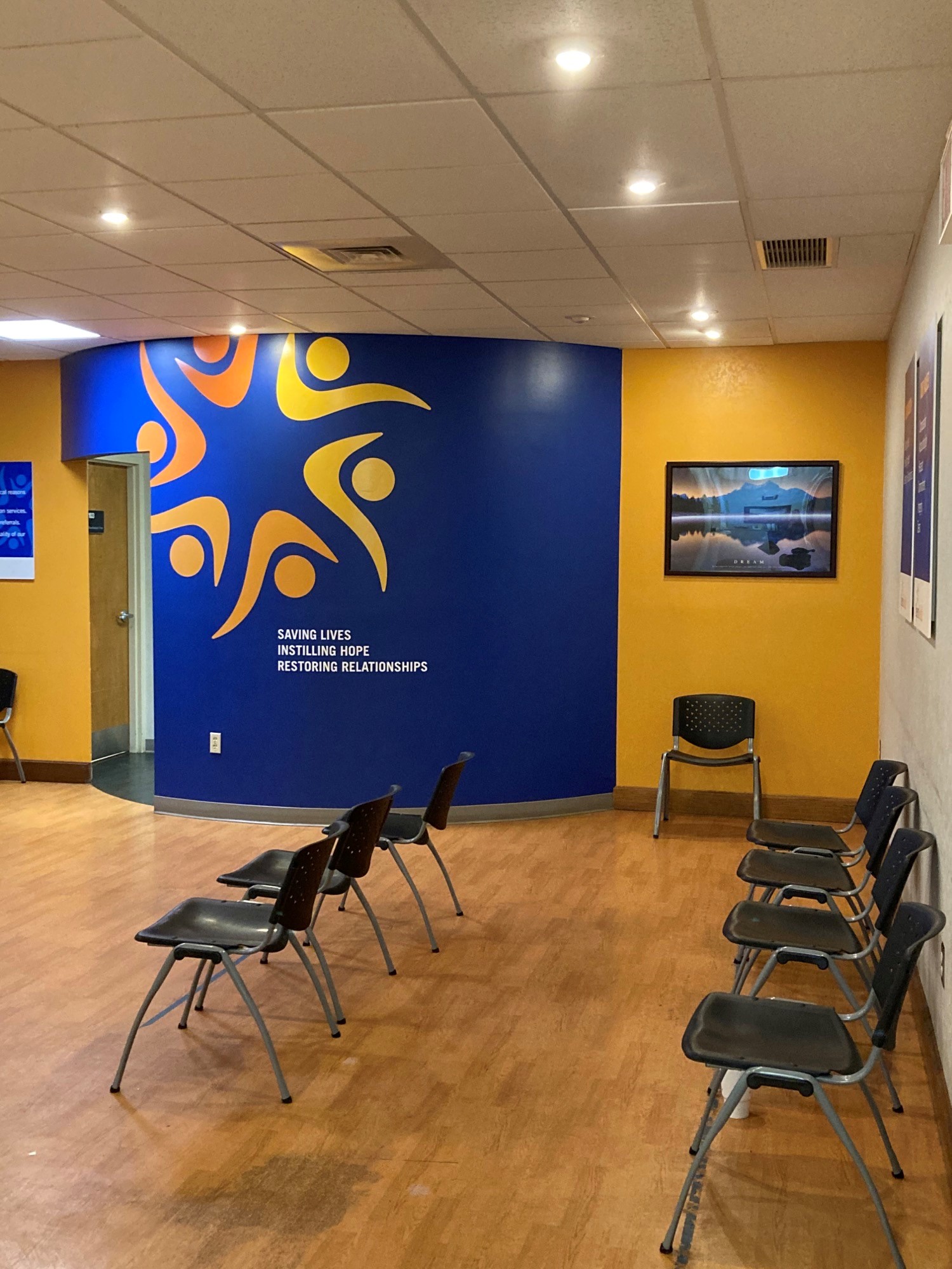 Interior room with blue and orange accent wall displaying facility branding, wooden flooring, rows of chairs, and mounted television screen