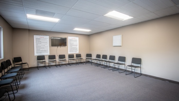 Interior meeting room with rows of black chairs in circular arrangement, cream walls, informational posters, television, lighting, and yellow door