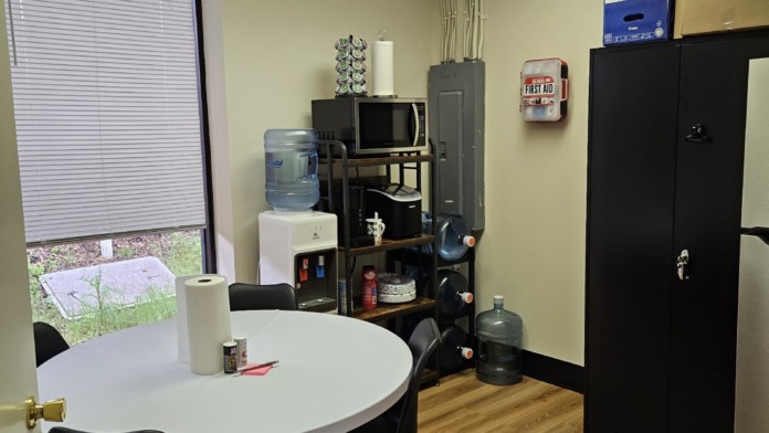 Interior community space with round table, black chairs, kitchenette area, television, and supplies storage.
