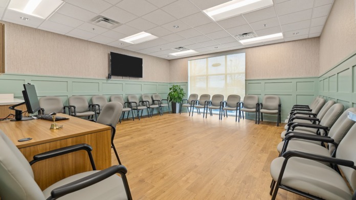 Interior group therapy room with multiple gray chairs in rows, green and white wall paneling, large TV monitor, and wooden flooring