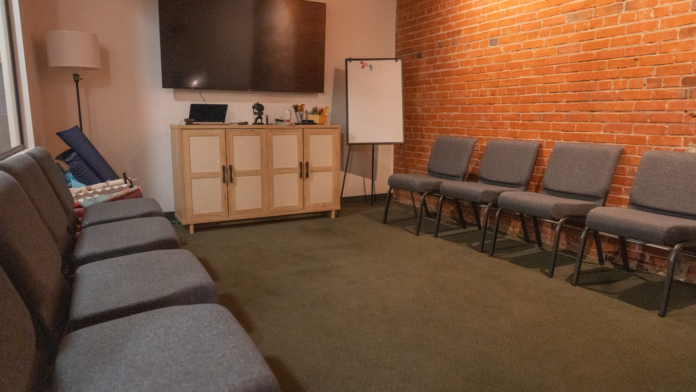 Interior room with gray upholstered chairs, television display, brick wall, whiteboard, and wooden cabinetry