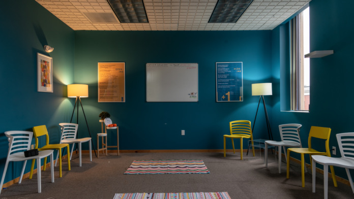Interior room with teal walls, yellow and white striped chairs in seating arrangement, wall-mounted posters, patterned rugs, table lamps, window