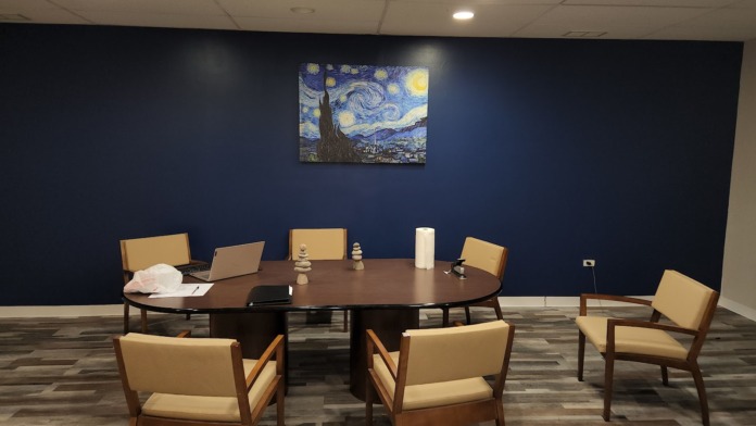 Interior therapy room with round dark wood table, tan upholstered chairs, blue accent wall, and artwork