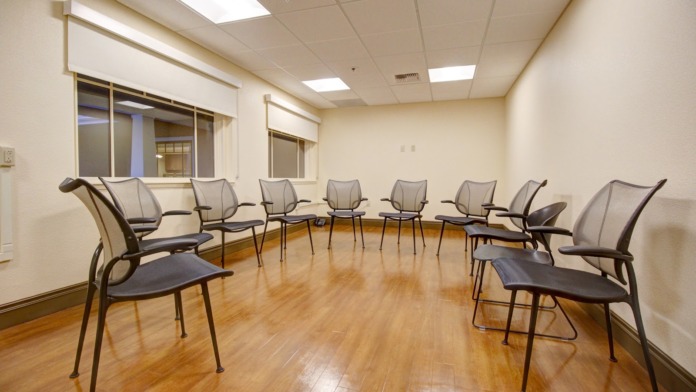 Interior therapy room with chairs arranged in circle formation, windows with blinds, wall-mounted equipment and natural lighting