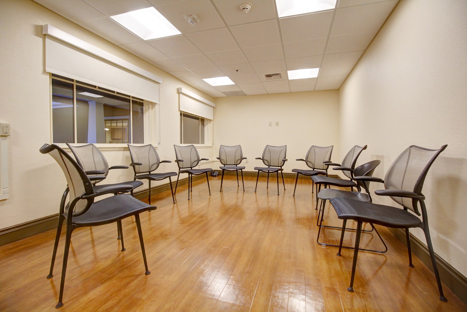 Interior therapy room with chairs arranged in circle formation, windows with blinds, wall-mounted equipment and natural lighting