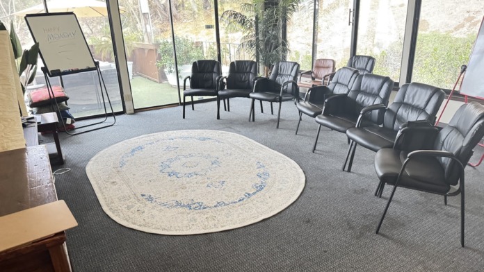 Interior circular gathering room with ring of black upholstered chairs, round rug, floor-to-ceiling glass walls, and exit signage.