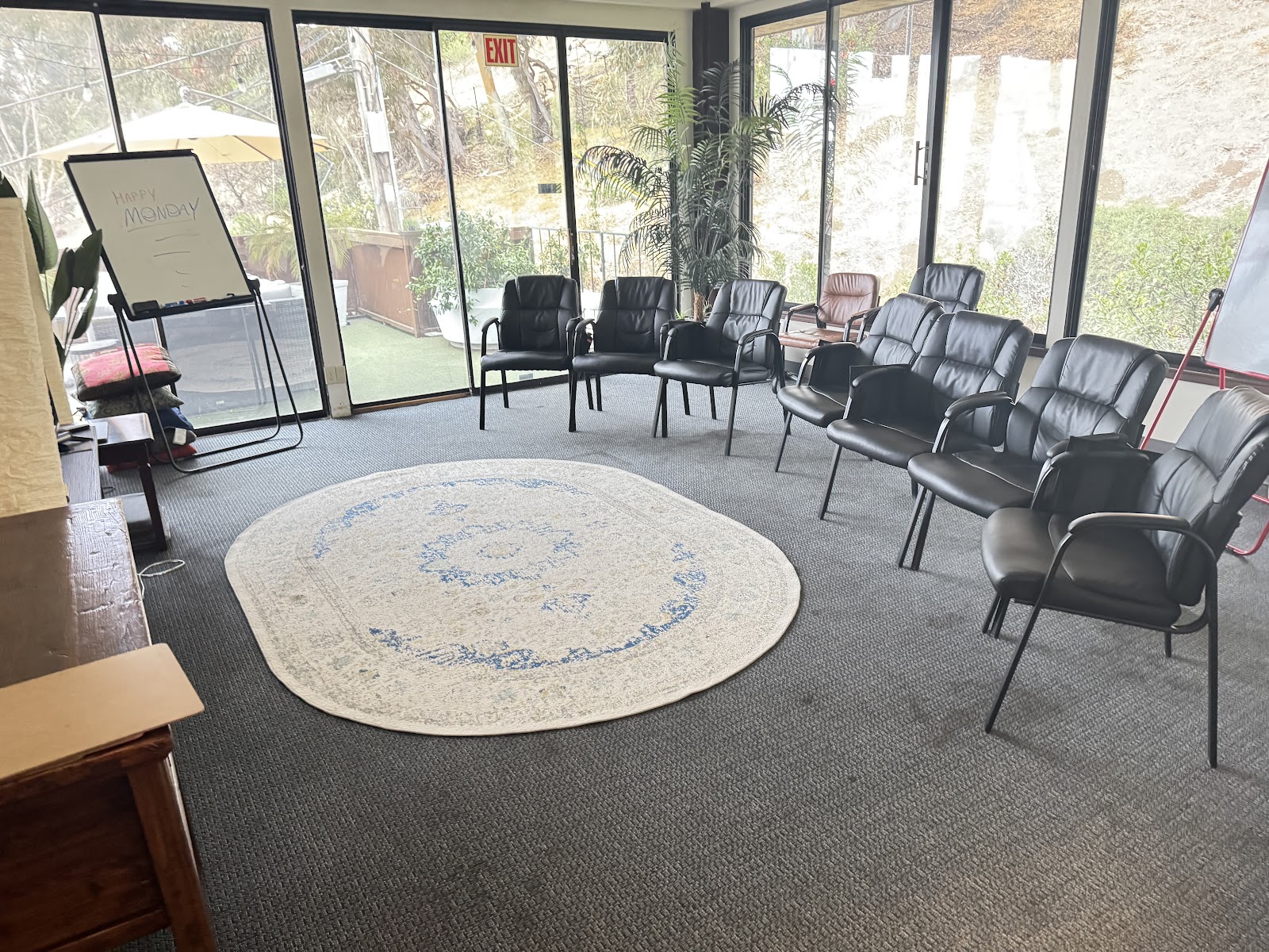 Interior circular gathering room with ring of black upholstered chairs, round rug, floor-to-ceiling glass walls, and exit signage.