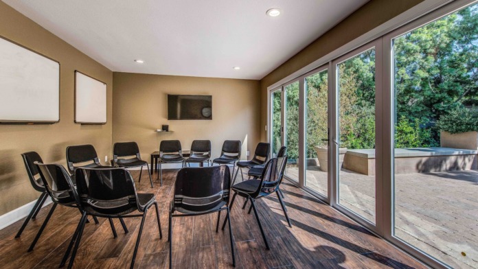 Interior therapy room with multiple black chairs arranged in circle formation, whiteboard, television, and glass doors opening to outdoor patio