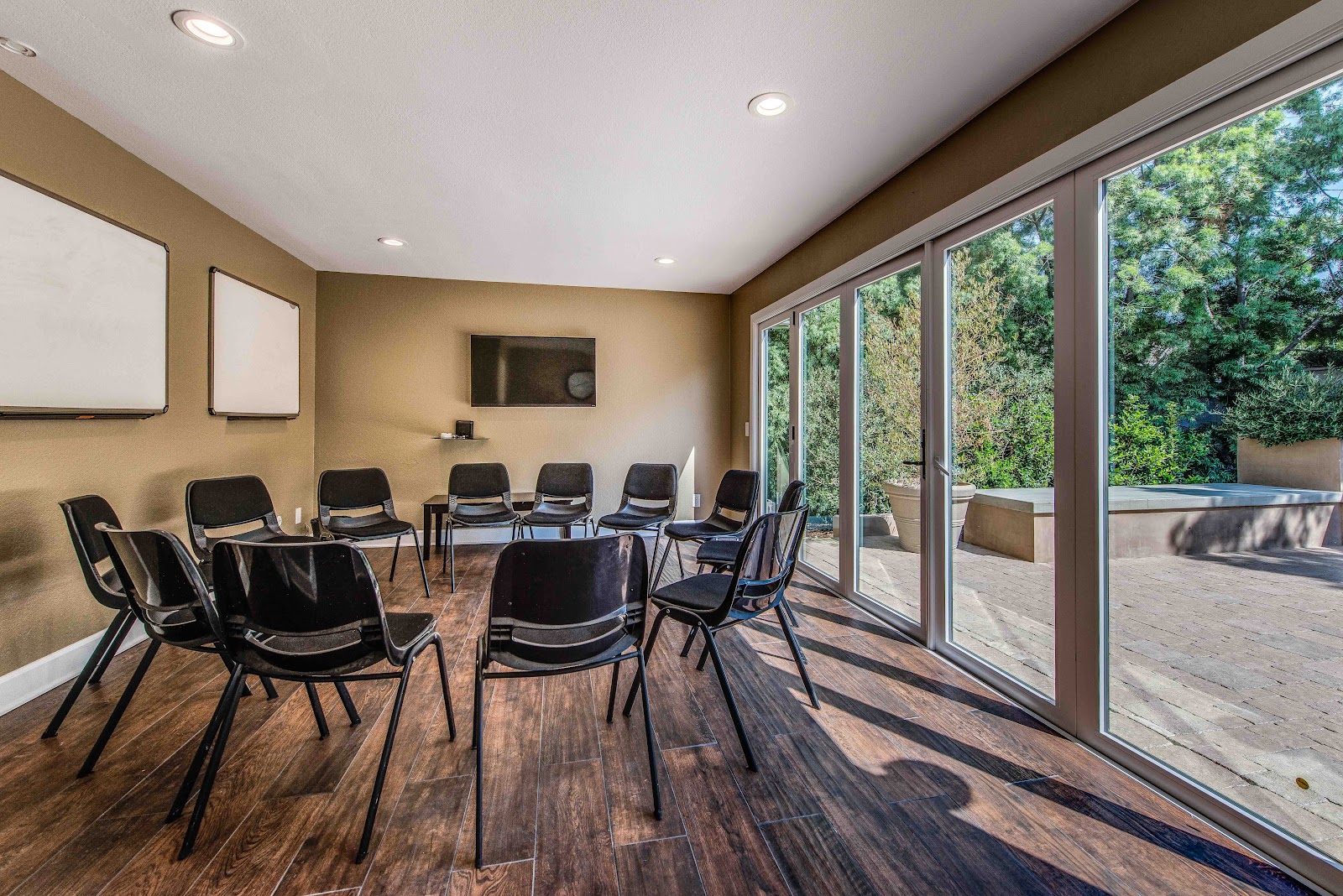Interior therapy room with multiple black chairs arranged in circle formation, whiteboard, television, and glass doors opening to outdoor patio
