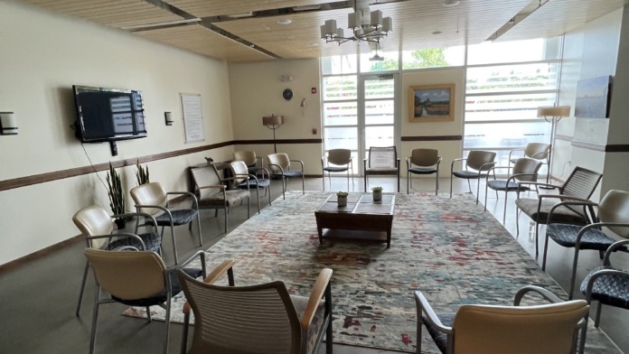Interior group therapy room with chairs arranged in circle, television on wall, large windows providing natural light.