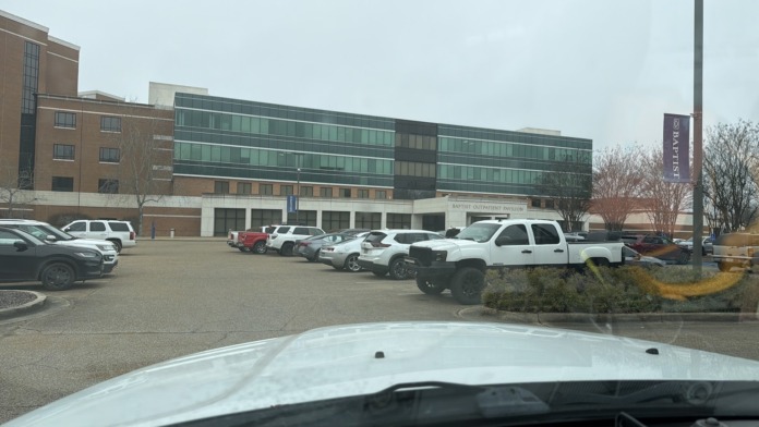 Exterior view of hospital complex showing brick and glass structures, parking area, and surrounding landscaping.