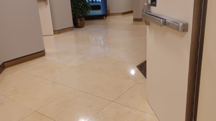 Interior hallway with polished tile flooring, beige walls, white doors, emergency signage, and display kiosk with decorative plant accents.