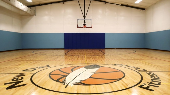 Interior gymnasium featuring wood flooring, basketball hoop, blue wall divider, and facility logo on court.