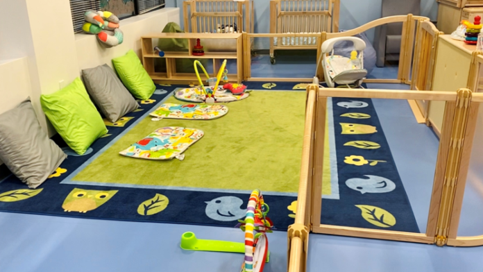 Interior playroom with colorful flooring, cushioned seating areas, storage cribs, and developmental toys for infants and toddlers