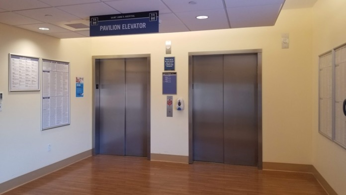 Interior elevator lobby with two elevator doors, signage, information boards, and wood flooring