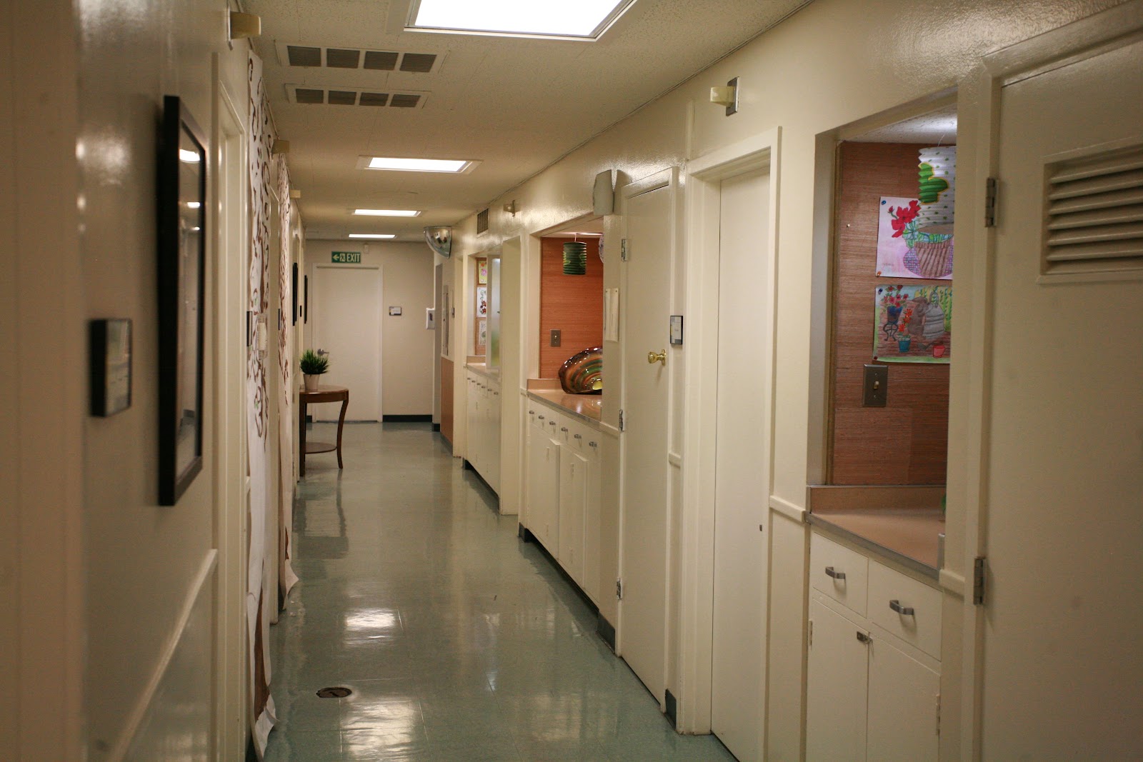 Interior hallway showing multiple patient room doors, white walls, medical signage, and emergency exit