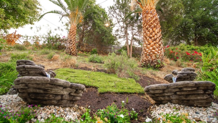 Outdoor garden featuring stone water fountains, palm trees, landscaped vegetation, and hillside terrain.