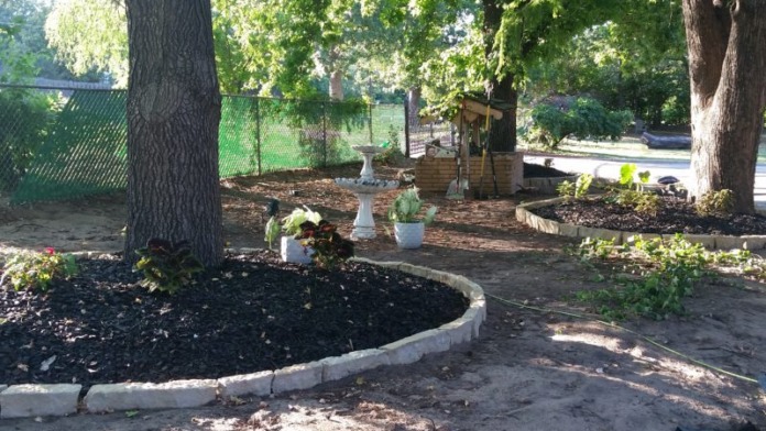 Outdoor garden area with mature trees, planted beds, circular mulch area, stone bird bath, and fencing