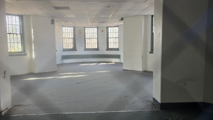 Interior open room with white ceiling, barred windows, columns, built-in seating, and industrial flooring.